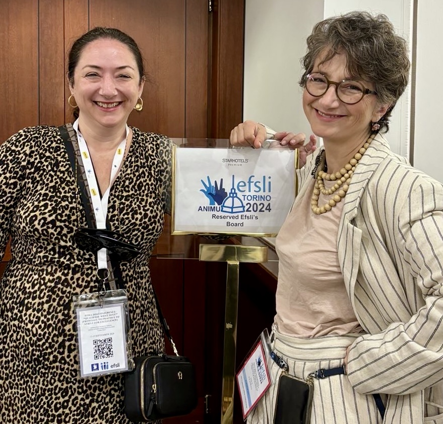 Oya und Ajda lächeln in die Kamera. Zwischen Ihnen ist ein Schild der Konferenz mit dem blauen Logo von ANIMU und "EFSLI Torino 2014"